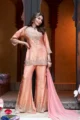 Peach Embroidered Shimmer Sharara Set: A woman posing in a premium 3-piece festive outfit featuring a shimmer-finish tunic with gold hand-embroidery, matching flared sharara pants, and a fluid georgette dupatta.